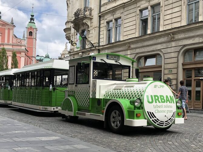 Green and white Urban electric tourist train driving through the cobblestone streets of Ljubljana old town. The pink Franciscan Church rises in the background as pedestrians walk nearby.