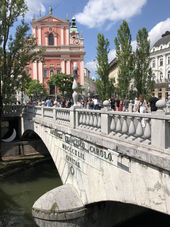 Crowds of people cross the Triple Bridge in Ljubljana Slovenia with the pink Franciscan Church of the Annunciation rising behind the stone balustrade on a sunny afternoon.