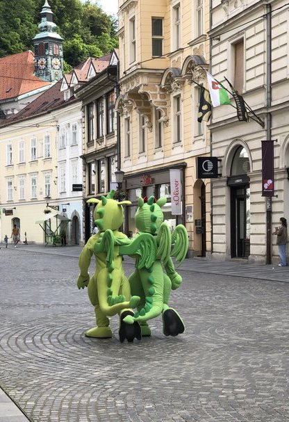 Two people in bright green dragon costumes walk arm-in-arm down a cobblestone street in Ljubljana’s old town, with historic buildings and the city's flag in the background. The playful scene captures the whimsical spirit of Ljubljana, known for its iconic dragon symbol.