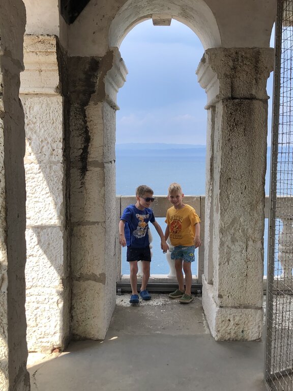 Two young boys stand under a stone archway overlooking the bright blue Adriatic Sea in Piran Slovenia. The weathered stone columns frame the ocean view behind them, highlighting the historic coastal architecture.