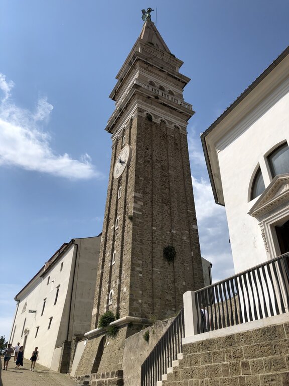 A tall historic bell tower rises against a blue sky in Piran Slovenia. The stone clock tower stands above surrounding buildings, emphasizing the town’s Venetian style architecture.