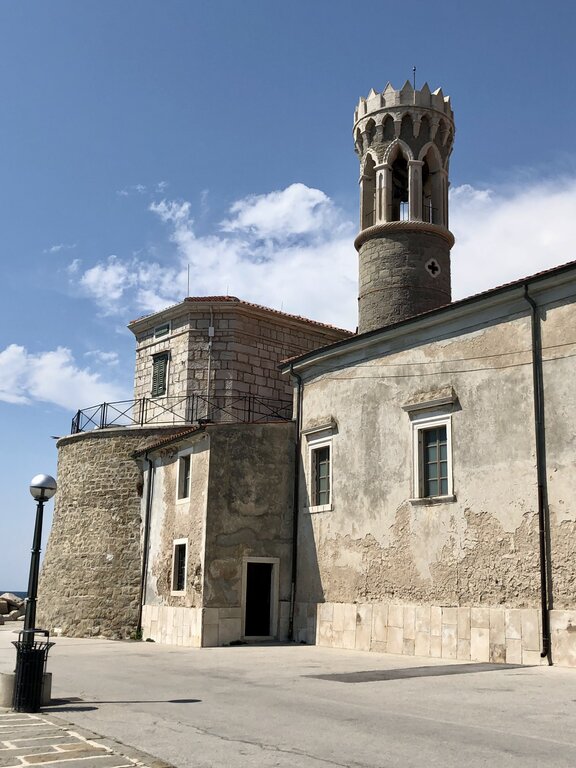 An old stone building with a small round turret and arched openings sits along a quiet street in Piran Slovenia. This is the lighthouse. The textured walls and pale stone exterior reflect the town’s historic coastal charm.