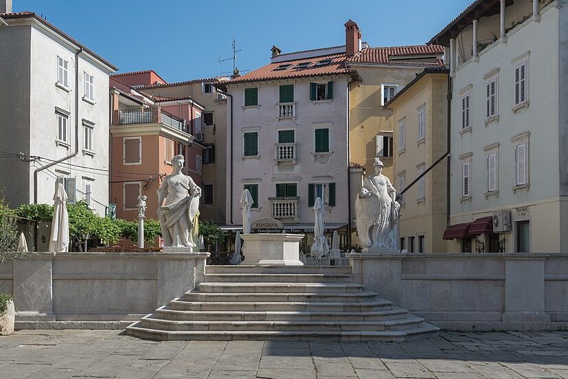 A stone fountain with classical statues stands in a quiet square in Piran Slovenia. Steps lead up to the raised platform, surrounded by pastel buildings with shutters and small balconies that reflect the town’s historic character.