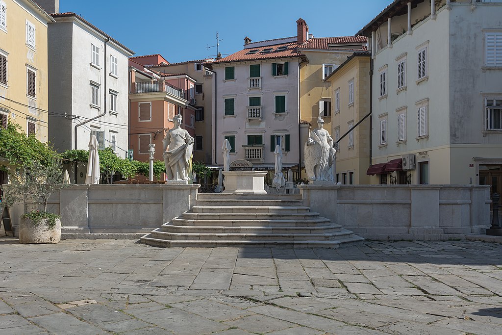 A stone fountain with classical statues stands in a quiet square in Piran Slovenia. Steps lead up to the raised platform, surrounded by pastel buildings with shutters and small balconies that reflect the town’s historic character.