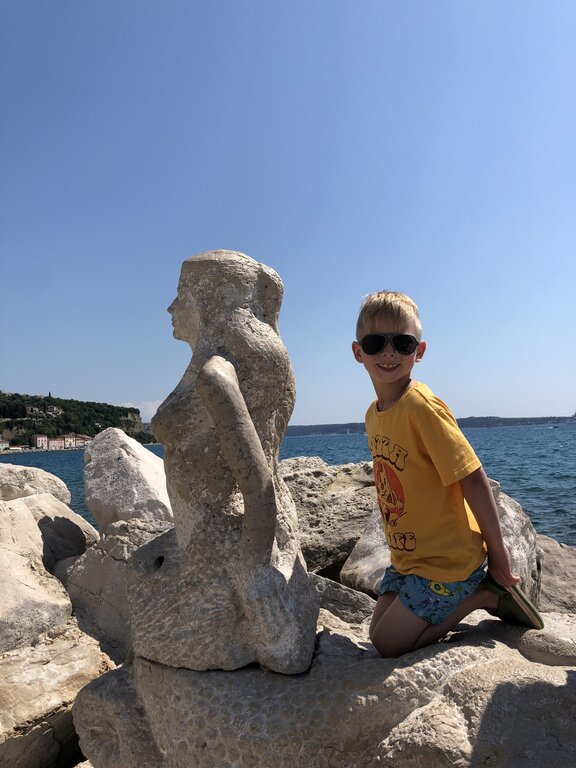 A young boy wearing sunglasses kneels beside a stone mermaid statue on rocky shoreline in Piran Slovenia. The calm blue sea behind them highlights the coastal setting.