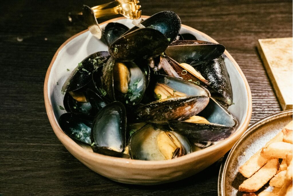 A bowl of steamed mussels in glossy black shells sits in a light ceramic dish, topped with chopped herbs and bathed in a buttery broth. The mussels are served on a dark wooden table with a side of golden fries partially visible.