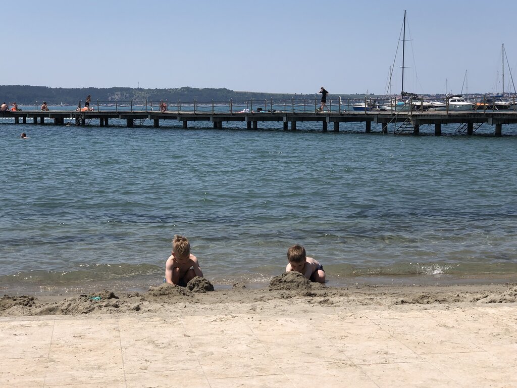 Two children build sand mounds at the edge of the water while a wooden pier stretches across the sea in Potoroz, Slovenia. Boats and swimmers in the distance emphasize the relaxed beach atmosphere.