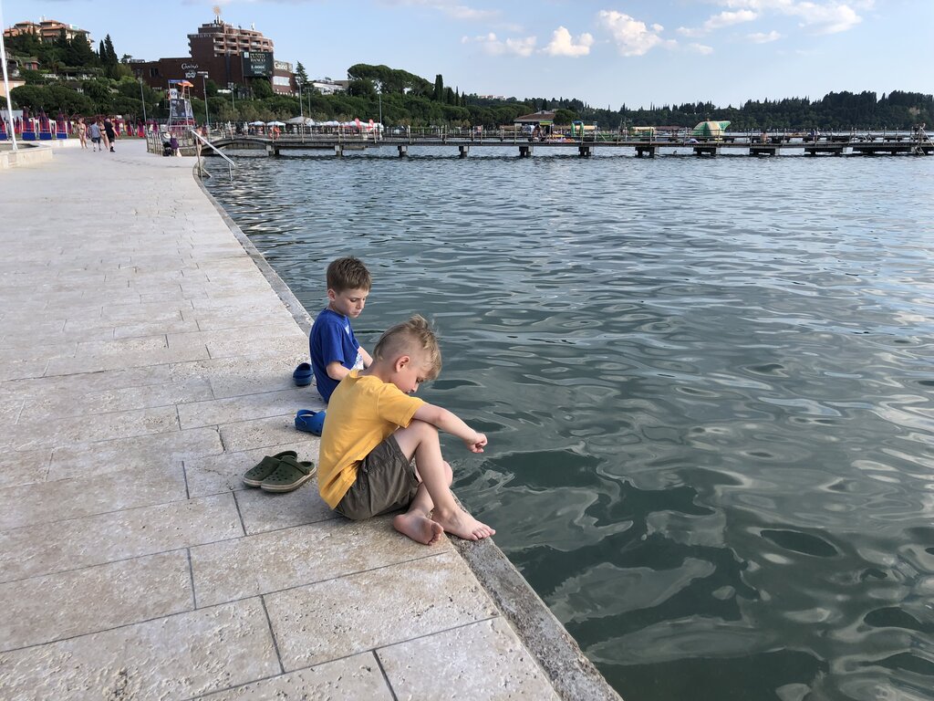 Two boys sit on a stone waterfront ledge dipping their feet into the water in Potoroz Slovenia. The calm harbour and nearby pier create a peaceful seaside moment.