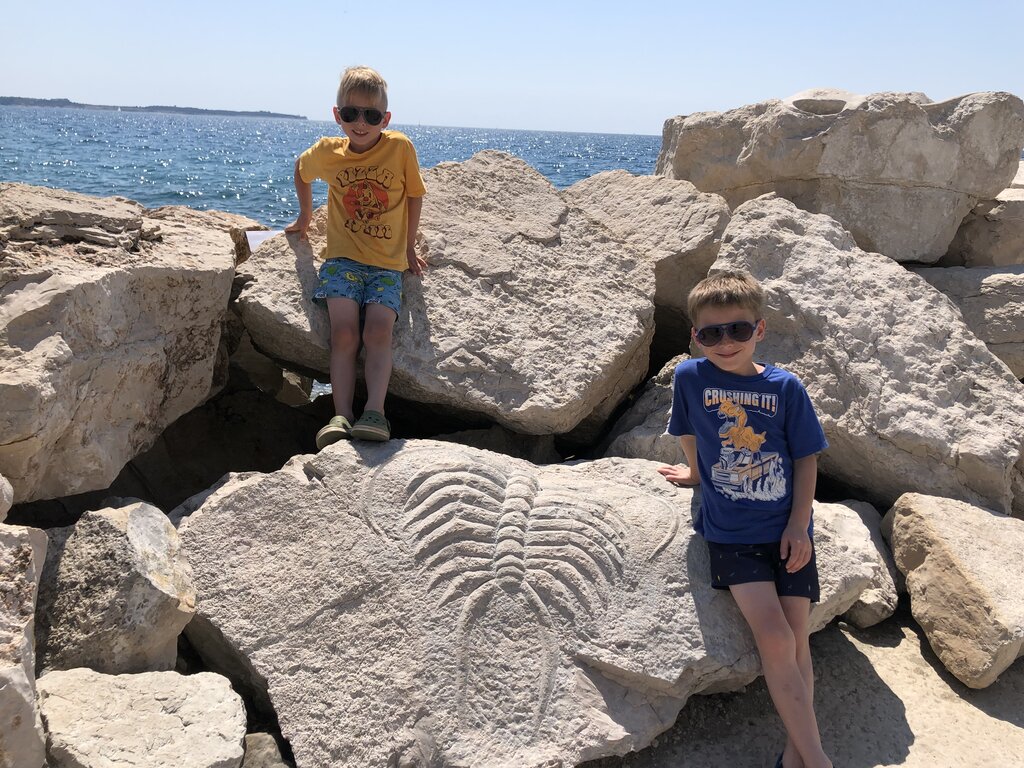 Two boys sit and stand on large seaside rocks beside a carved stone relief near the Adriatic Sea in Piran Slovenia. The bright sunlight and sparkling water create a playful summer atmosphere.