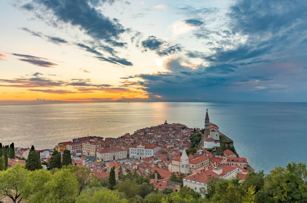 An elevated sunset view of Piran Slovenia shows red tiled rooftops stretching toward the Adriatic Sea. The church and bell tower sit prominently on the peninsula as golden light breaks through dramatic clouds over the water.