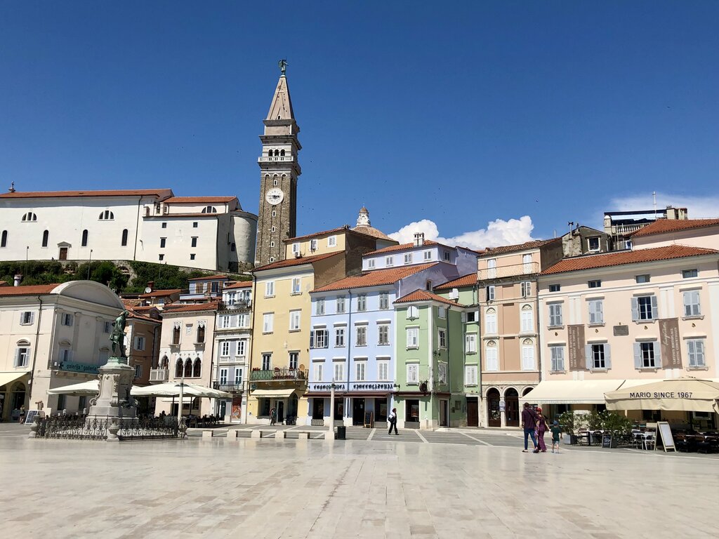 Colourful pastel buildings line the wide main square, Tartini Square, in Piran Slovenia with a tall bell tower rising in the background. The open plaza and bright facades showcase the lively historic town center.