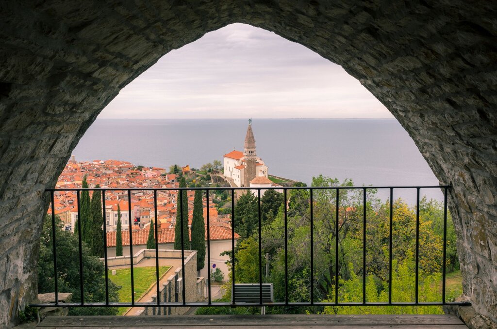 A scenic view of Piran Slovenia is framed by a stone archway and iron railing overlooking the town and the Adriatic Sea. The bell tower rises above red rooftops while greenery fills the foreground.