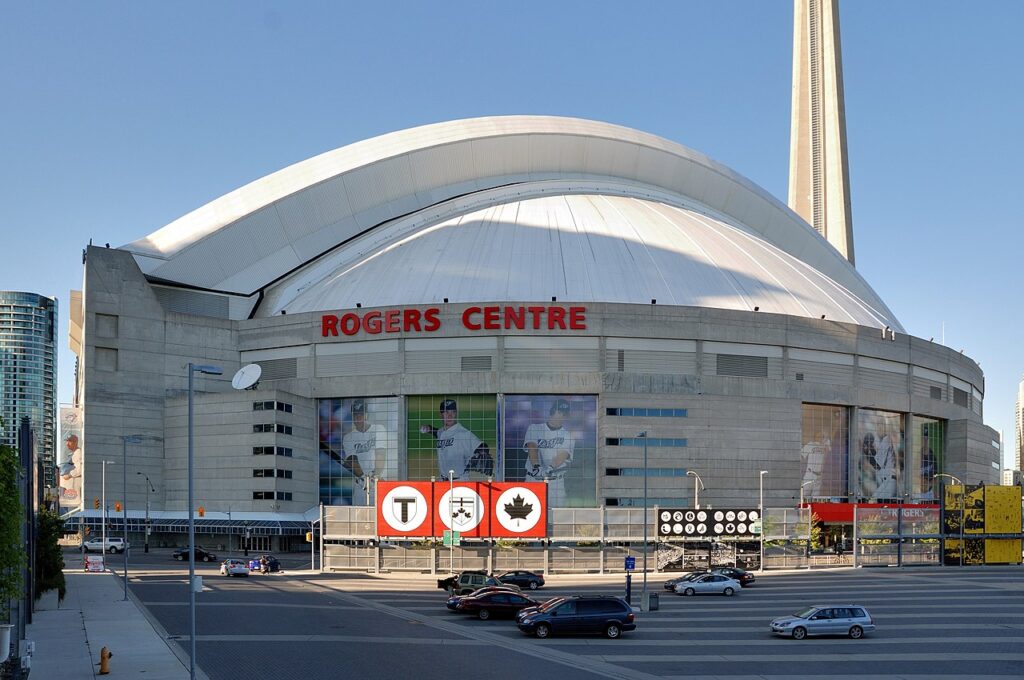 The exterior of Rogers Centre in downtown Toronto, with its distinctive retractable dome roof partially open, large Toronto Blue Jays player murals on the facade, and the CN Tower rising behind it against a clear blue sky.