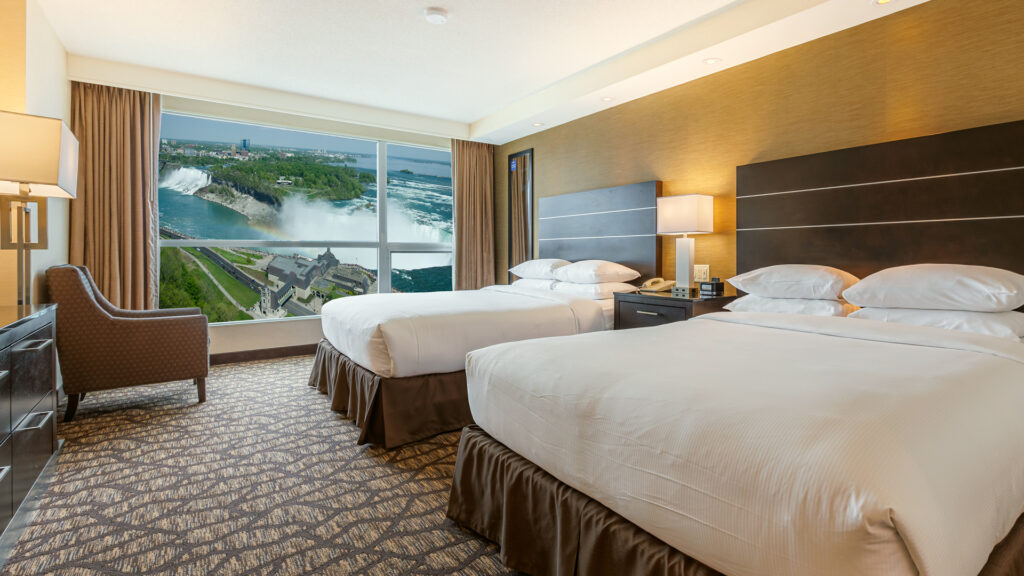 A double queen hotel room at Embassy Suites Niagara Falls with warm wood accents and a large window offering a high-altitude view of Horseshoe Falls, the Niagara River, and the surrounding parkland.