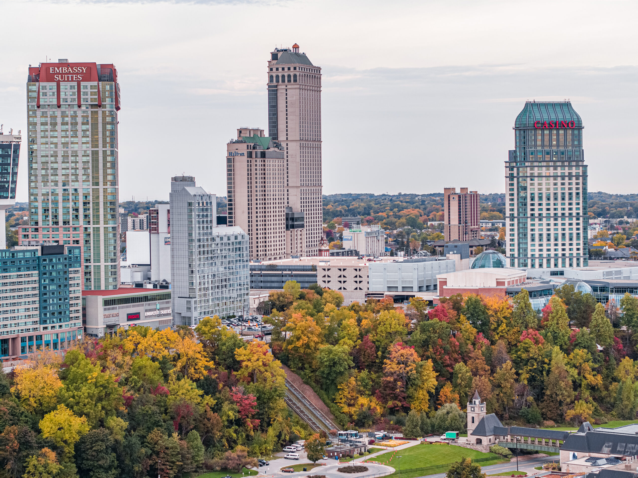 An aerial autumn view of the Niagara Falls, Ontario hotel district, featuring the Embassy Suites tower, Hilton, and Casino Niagara rising above a colourful canopy of red, orange, and yellow fall foliage in Queen Victoria Park.