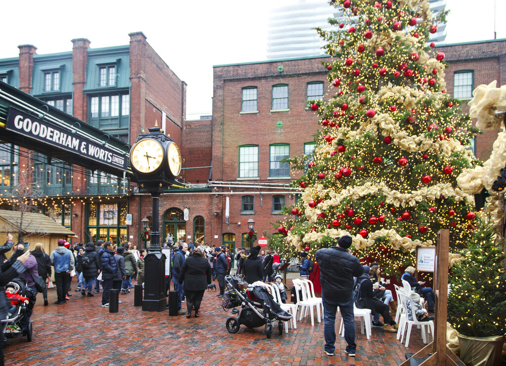 A large decorated Christmas tree covered in lights red ornaments and gold ribbon stands in a busy courtyard at Toronto’s Distillery District. Families gather around the tree near a sign reading "Gooderham & Worts Limited" while people stroll along the brick streets during the holiday market.