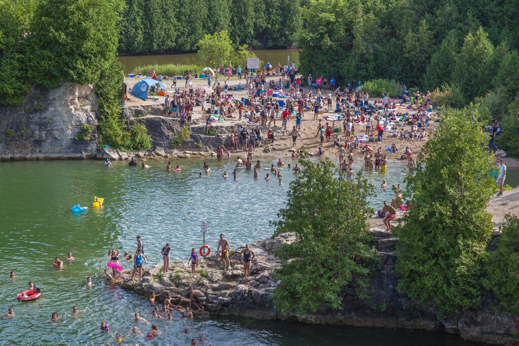 A busy summer day at Elora Quarry Conservation Area, with crowds of swimmers and sunbathers packed along the rocky limestone shoreline of the clear green quarry pool, surrounded by dense trees.