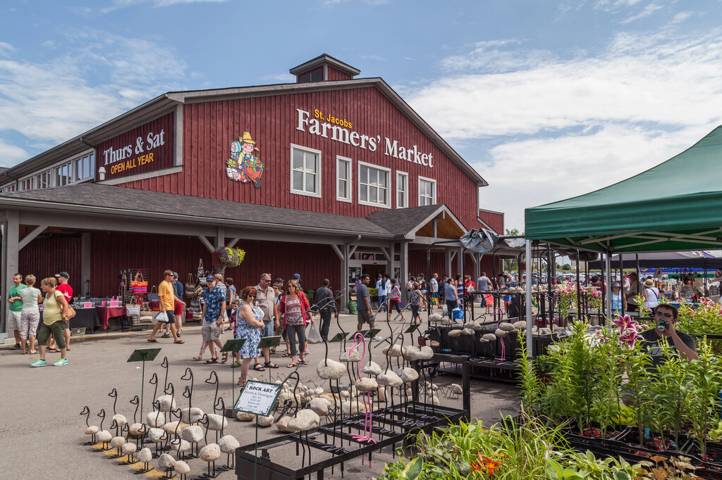 Crowds of shoppers browse outdoor vendor stalls in front of the red barn-style St. Jacobs Farmers' Market building in Ontario, with signage noting it's open Thursdays and Saturdays all year.