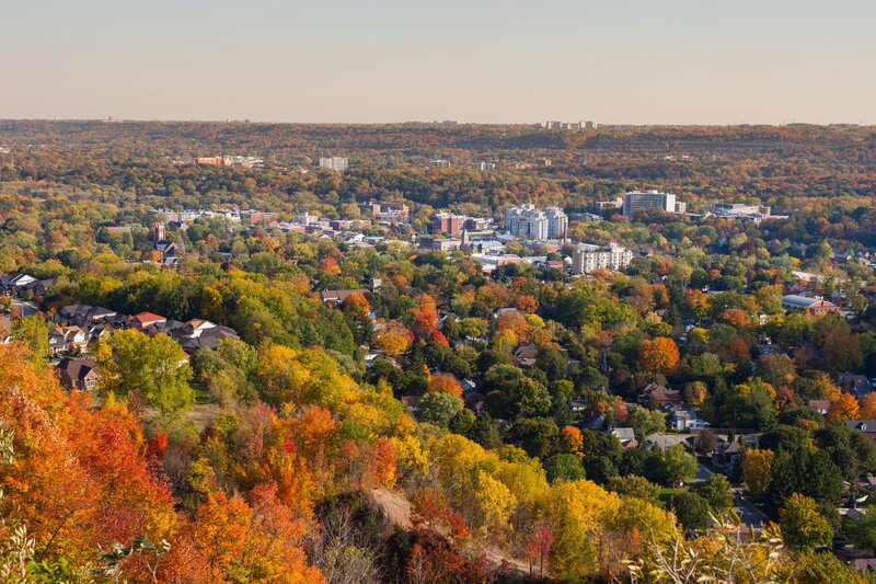 An elevated autumn view over the town of Dundas, Ontario, with vibrant red, orange, and green fall foliage in the foreground giving way to a mix of residential neighbourhoods and low-rise downtown buildings, backed by the Niagara Escarpment ridge on the horizon.