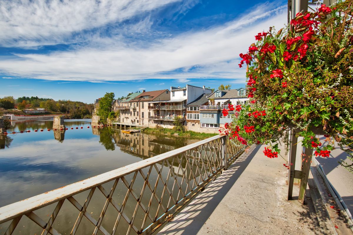 Riverside walkway in Elora Ontario with a metal railing beside calm water reflecting historic buildings with balconies along the opposite bank. Red flowers hang in the foreground on the right under a bright blue sky with scattered clouds.