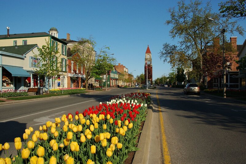 The main street of Niagara-on-the-Lake in spring, with a colourful median of yellow, red, and white tulips lining the road, historic storefronts and mature trees on either side, and the town's iconic red-brick clock tower visible in the distance.