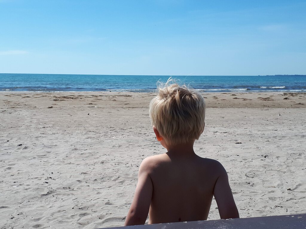 Young child with short blonde hair sits on a sandy beach facing the ocean with gentle waves rolling in under a clear blue sky. The wide open shoreline stretches ahead with no other people nearby creating a quiet peaceful scene.