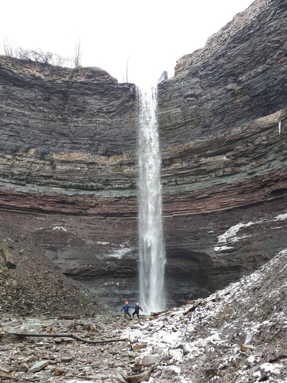 Tall waterfall plunging from the top of a layered rock cliff into a rocky basin below at Devil's Punchbowl in Hamilton ONtario. Two young children stand at the bottom showing the height of the narrow waterfall.