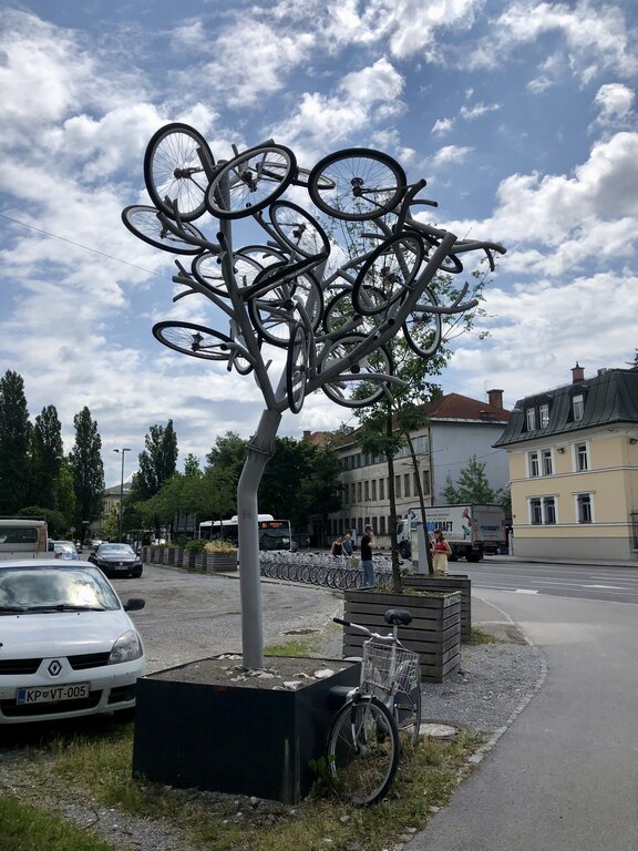 Metal sculpture shaped like a tree with multiple bicycle frames and wheels forming the branches. The bike tree stands on a city sidewalk next to parked cars and residential buildings under a partly cloudy sky, highlighting a creative urban art installation made from recycled bicycles.