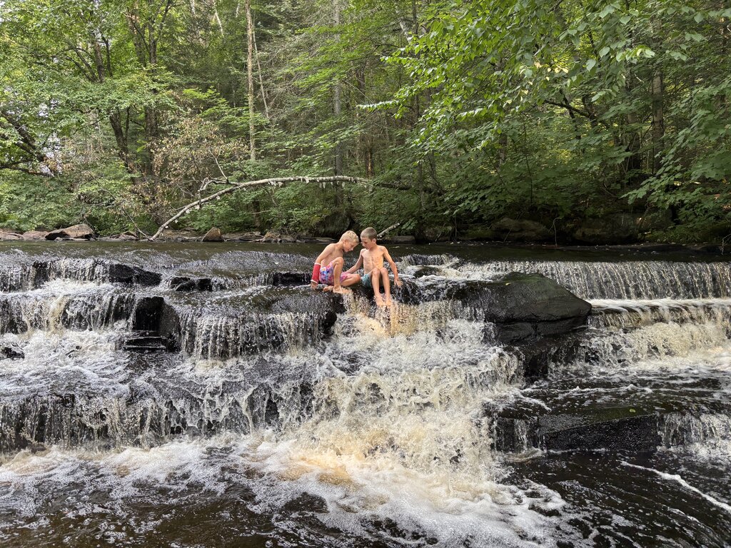 Two children sit on rocks at the top of a small cascading waterfall in a forested area in Muskoka, Ontario. Water flows around them as they look down at the stream below, surrounded by dense greenery.