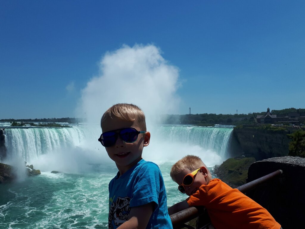 Two children wearing sunglasses stand at a railing with Horseshoe Falls crashing behind them at Niagara Falls. Mist rises high above the powerful waterfall as the kids look toward the rushing water.