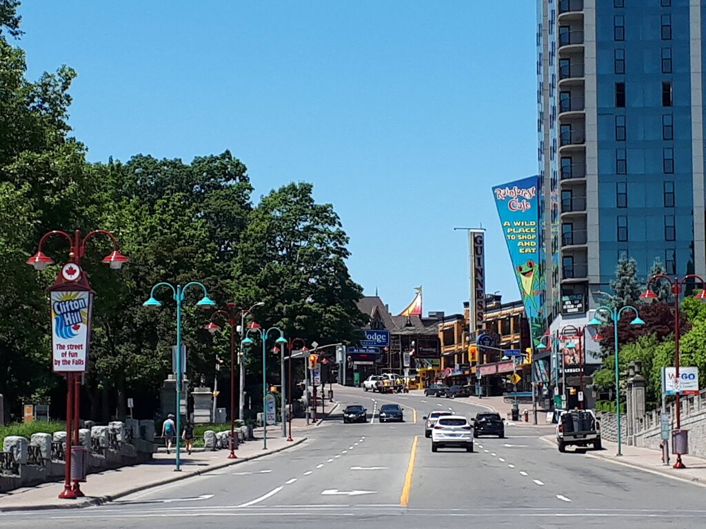 A street in Niagara Falls lined with colorful signs and lampposts including one that reads "Clifton Hill The street of fun by the Falls". Cars drive up the road toward restaurants attractions and hotels.