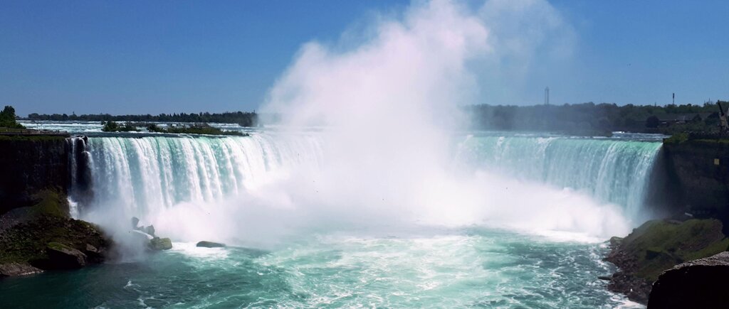 Horseshoe Falls at Niagara Falls crashes into the river below with thick mist rising from the powerful waterfall. The wide curved falls stretch across the horizon under a bright sky.
