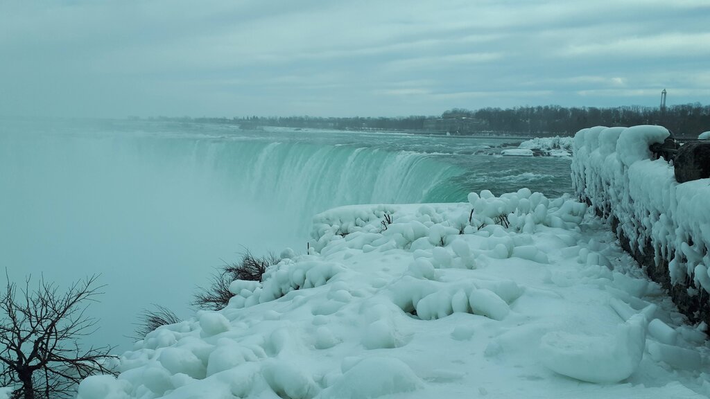 The edge of Niagara Falls in winter with thick ice and snow covering the rocks beside the rushing water. Mist rises above the frozen landscape as the falls plunge into the river below.