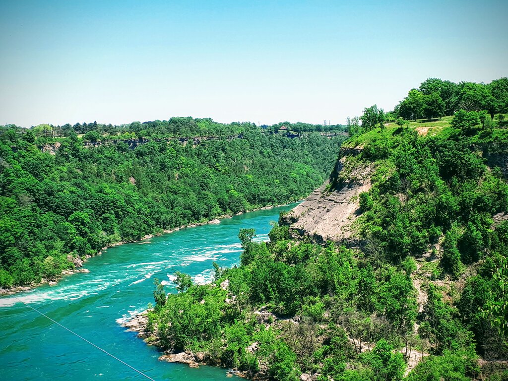 The Niagara River winds through a deep green gorge with steep rocky cliffs on either side. Trees cover the hills surrounding the bright blue water below.