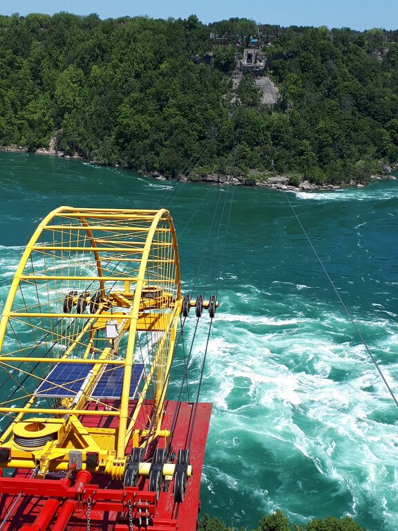 A bright yellow whirlpool aero car cable system hangs above the swirling turquoise water of the Niagara Whirlpool. The metal structure and cables stretch across the river between forested cliffs.