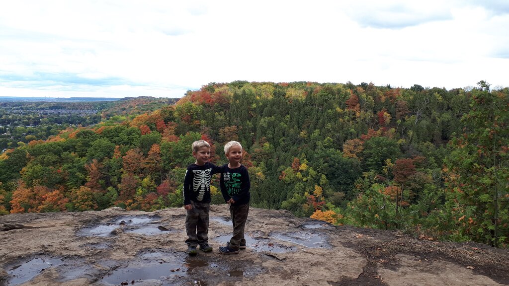 Two young children stand on a rocky cliff at Beamer Conservation area amid the fall colours of the Niagara escarpment . The wide scenic view shows red orange and green autumn trees stretching across the valley.