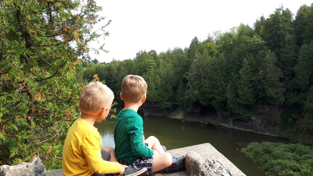 Two young children sit on a rock ledge overlooking a calm river surrounded by dense green forest with a cave visible across the water.