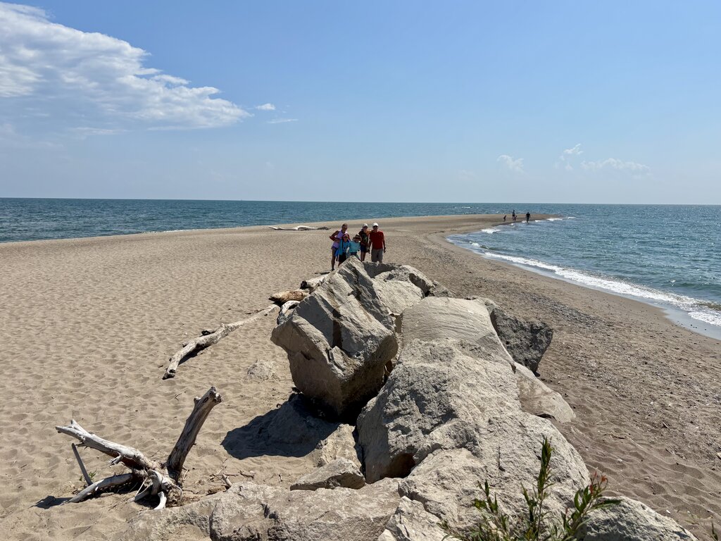 a group of people stand on large rocks at the tip of a sandy point at Point Pelee, the southernmost point in Canada, with water on both sides and a clear blue sky overhead.