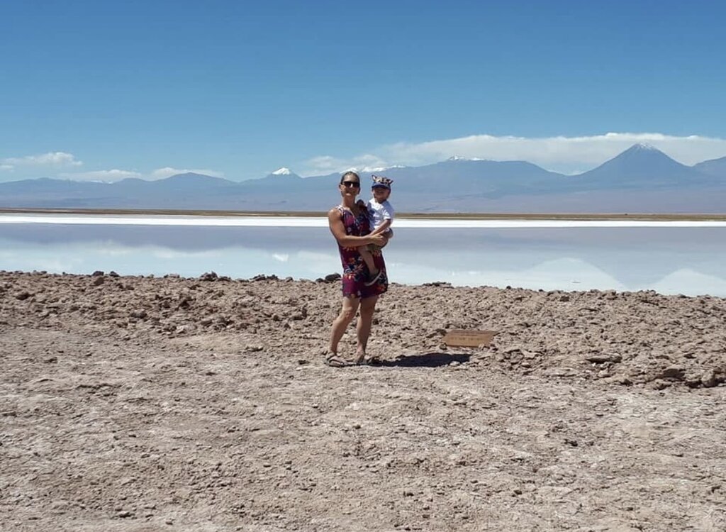 Woman holding a young child stands on a rocky shoreline of the salt flats in Chile with calm reflective water and distant mountains under a clear blue sky.