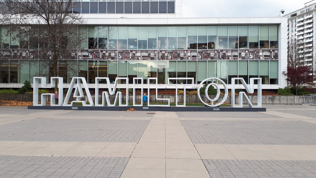 Large metal letters spelling "HAMILTON" stand in front of a modern building at City Hall. A small child stands between the letters in the open plaza.