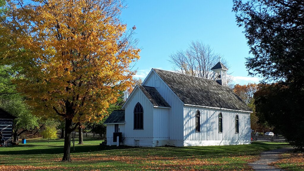 A small historic white wooden chapel with arched windows and a bell tower at Ball's Falls Conservation Area in Niagara, surrounded by autumn foliage, fallen leaves, and a large tree with golden-orange leaves under a clear blue sky.