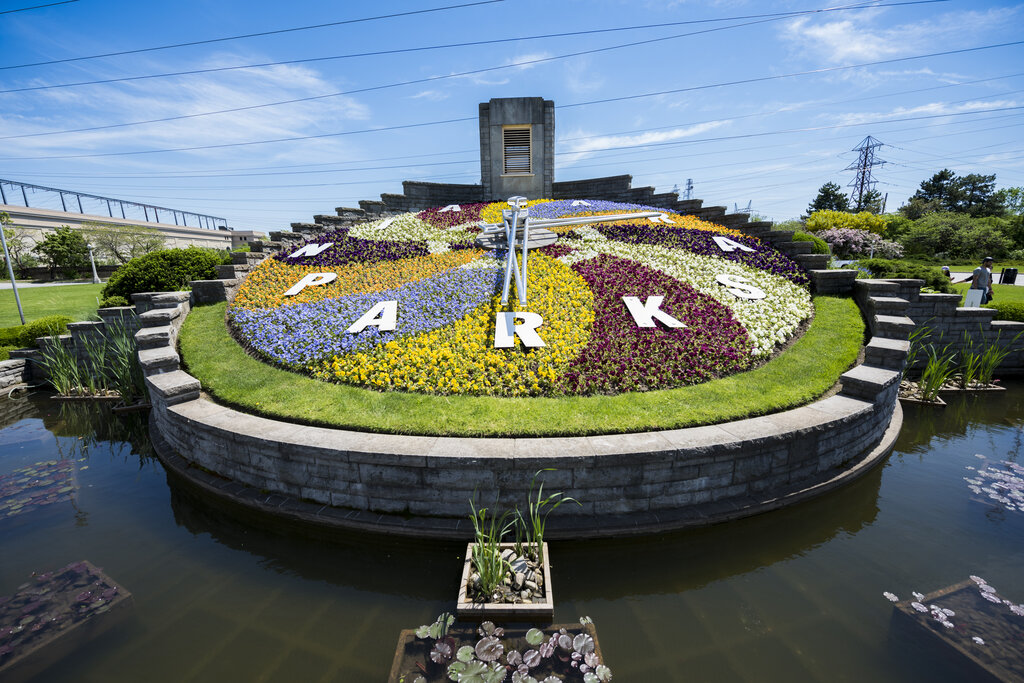 Large circular floral clock made of colorful flowers and green grass displays the word "PARK" with white letters and metal clock hands in a landscaped garden near Niagara Falls. The flower beds form bright sections of yellow purple white and red around the clock face surrounded by a shallow reflecting pond.
