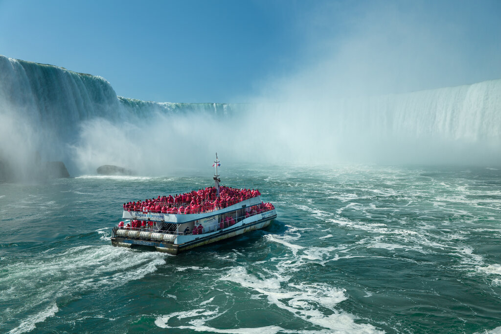Tour boat filled with visitors in red ponchos approaching the base of Niagara Falls as mist rises from the massive waterfall and churns across the turquoise river. The boat heads toward the curved wall of Niagara Falls while passengers gather on the upper deck to experience the spray and power of the water.