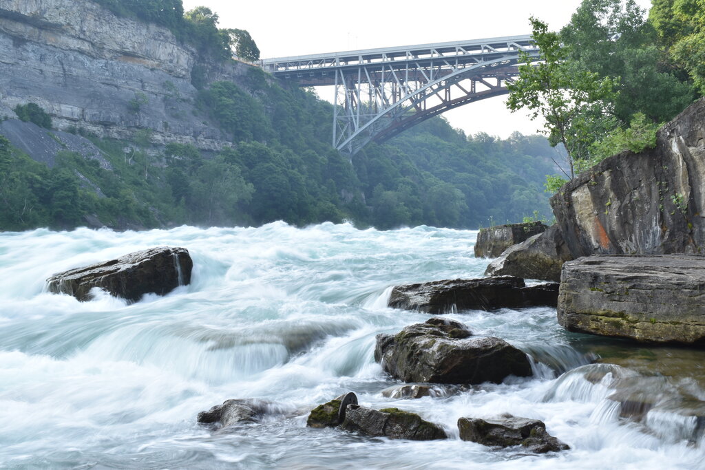 Fast moving whitewater rapids of the Niagara River rush past large rocks in the gorge below a tall steel arch bridge surrounded by green cliffs near Niagara Falls. The turbulent water churns through the narrow gorge with mist rising in the distance.
