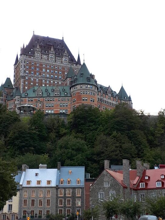 A large castle-like hotel with green rooftops rises above colourful historic stone houses nestled at the base of a hill covered in trees. This is the Chateau Frontenac in Quebec City