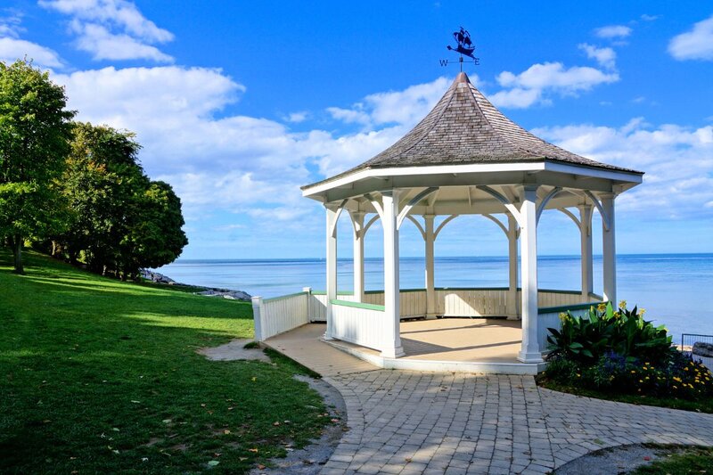A white Victorian-style gazebo with a weather vane on top, situated on a brick pathway overlooking the calm blue waters of Lake Ontario in Niagara-on-the-Lake, surrounded by green lawns and trees under a bright blue sky with scattered clouds.