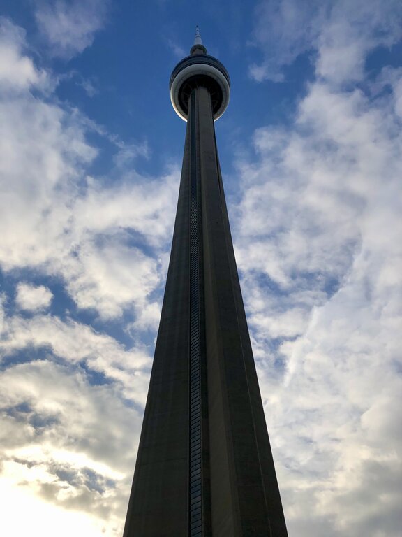 The CN Tower photographed from directly below with the concrete tower stretching high into a partly cloudy sky. The circular observation deck sits near the top creating a dramatic upward view of Toronto’s most recognizable landmark.