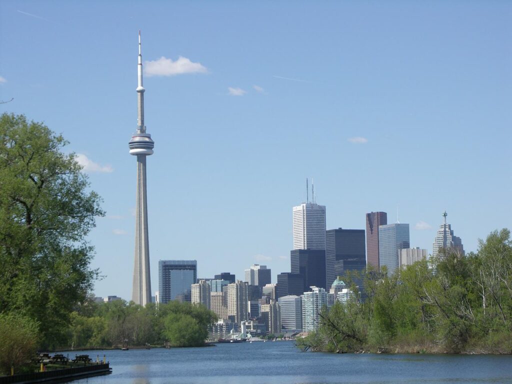 The CN Tower rising above the Toronto skyline on a clear day viewed across calm water and green trees. The tall tower stands beside downtown skyscrapers creating an iconic city view often seen when visiting Toronto with kids.