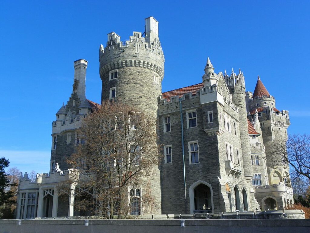 Casa Loma castle in Toronto, a grand early 20th-century Gothic Revival mansion featuring grey stone towers, turrets with red-tiled roofs, arched entryways, and ornate battlements, photographed on a clear sunny day with bare winter trees in the foreground.