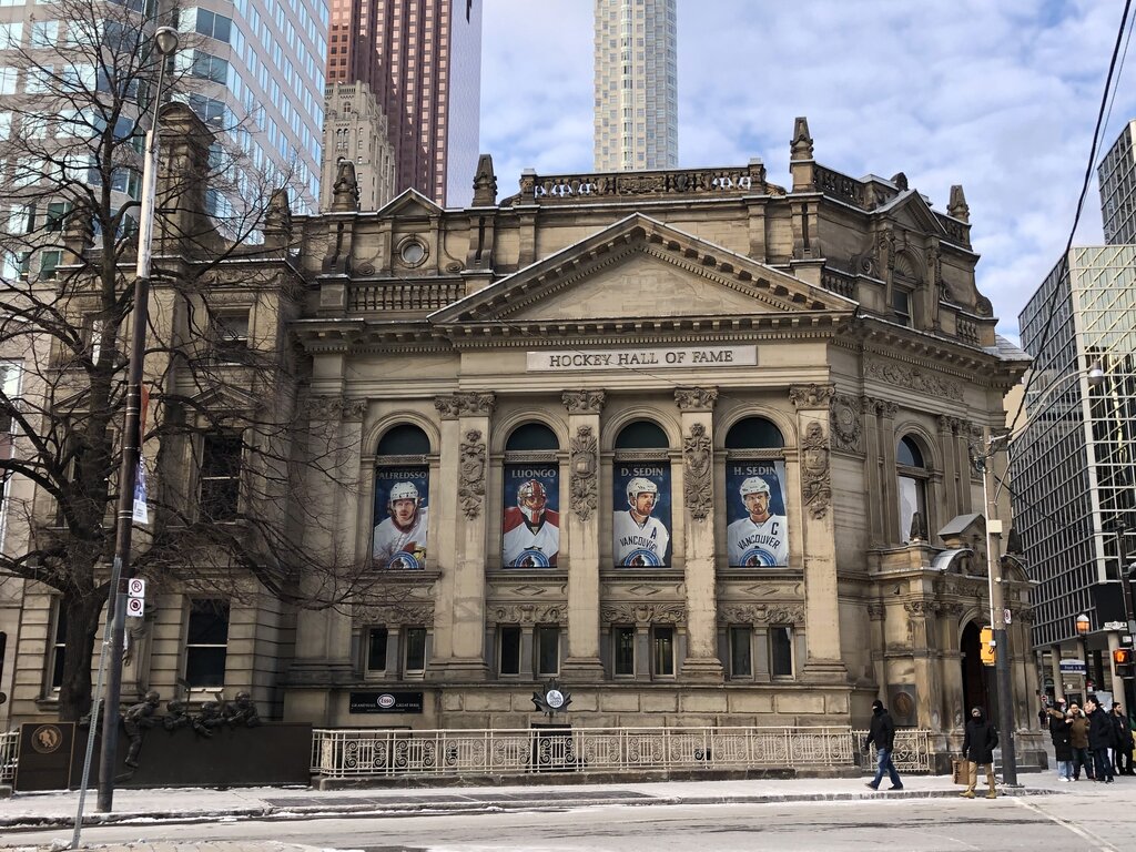 Historic stone building of the Hockey Hall of Fame in downtown Toronto with tall city skyscrapers behind it. Banners of hockey players hang between columns beneath the carved sign that reads "Hockey Hall of Fame".